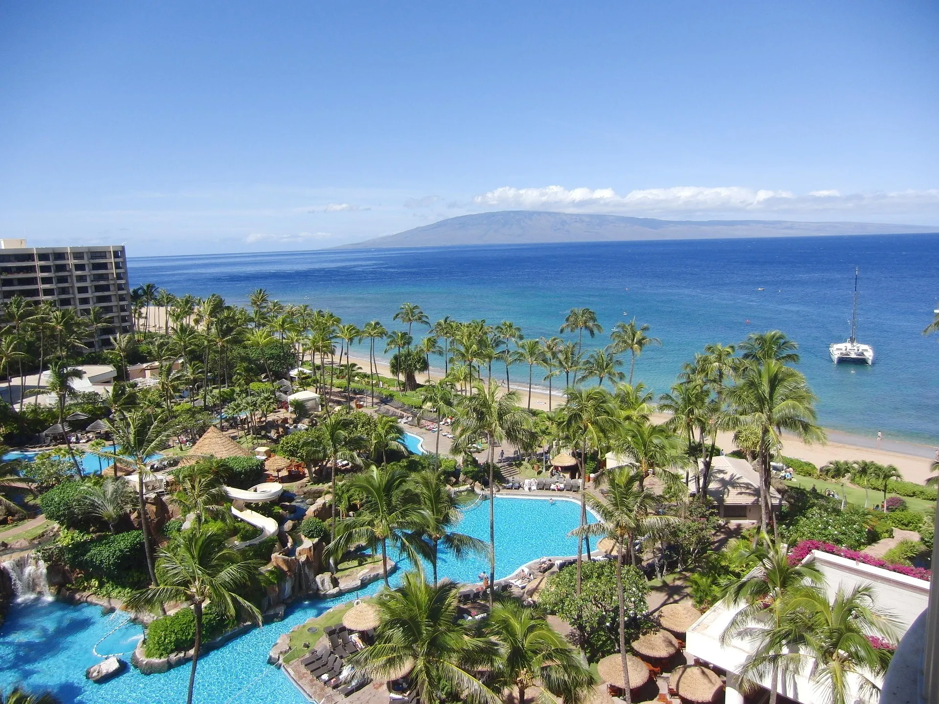 An elevated, panoramic view of a luxury resort in Maui, Hawaii, featuring winding turquoise swimming pools, lush palm trees, and a white sand beach bordering the deep blue Pacific Ocean with a catamaran at anchor and the island of Lanai on the horizon.