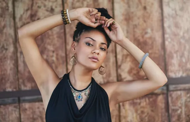 Hot girl with short, dark curly hair, wearing a black top, layered necklaces with a prominent blue pendant, large hoop earrings, and multiple bracelets, poses with her arms raised and hands near her head against a wooden wall.