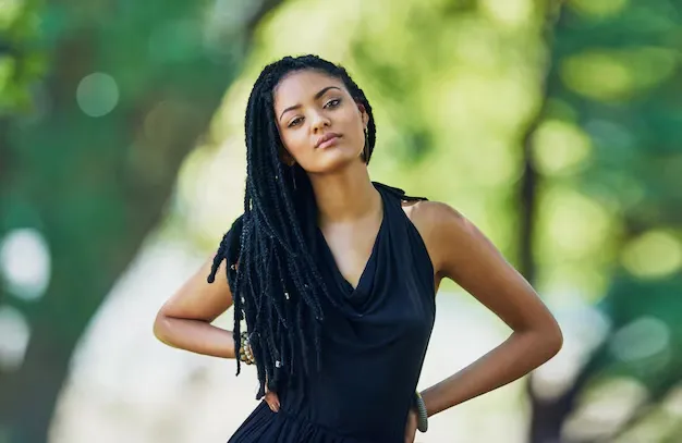 Hot girl with long, dark dreadlocks, wearing a black V-neck top, stands confidently with her hands on her hips, against a blurred green outdoor background.