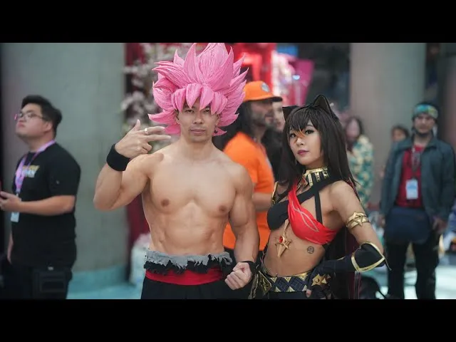 A male cosplayer dressed as Goku Black in Super Saiyan Rosé form (pink spiky hair) poses alongside a female cosplayer in an elaborate fantasy costume at an anime convention.