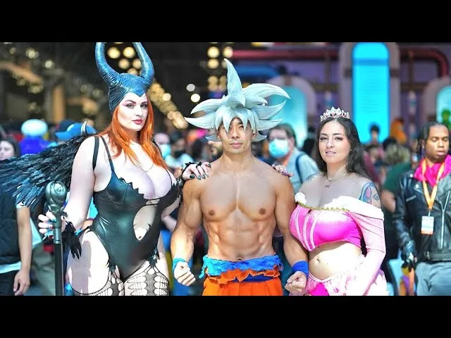 Three cosplayers pose at a convention, featuring a male cosplayer as Ultra Instinct Goku flanked by female cosplayers dressed as a horned dark fairy and a princess in a pink gown.