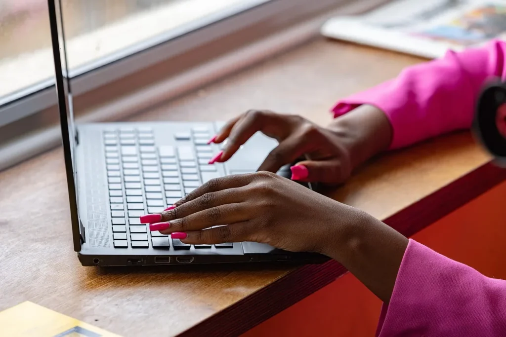 Close-up of a Black woman's hands with bright pink nails typing on a laptop, emphasizing a personalized and active online presence.