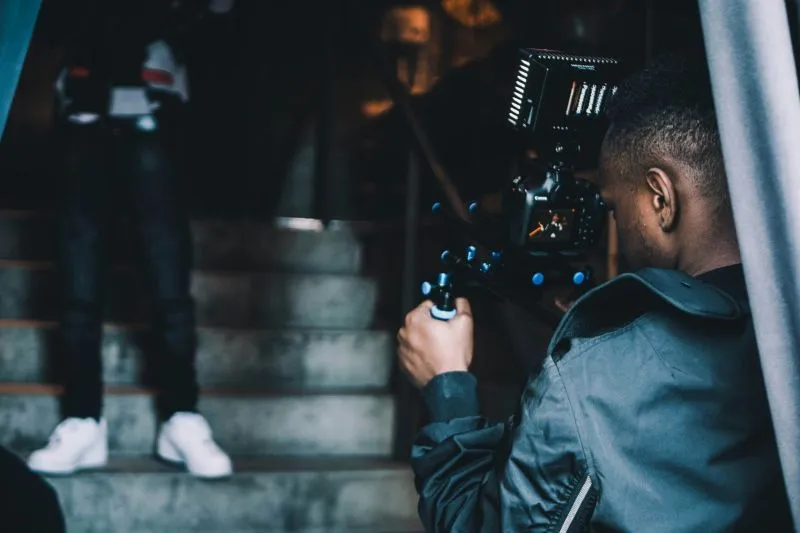 A person holding a professional video camera, filming a subject (partially visible) walking up a flight of stairs in a dimly lit, indoor setting.