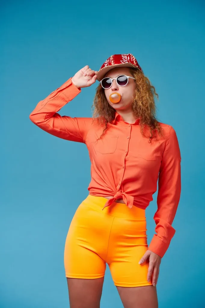 A woman with curly hair, wearing a red cap, sunglasses, an orange button-up shirt, and orange shorts, blowing a large bablbabldabl gum.