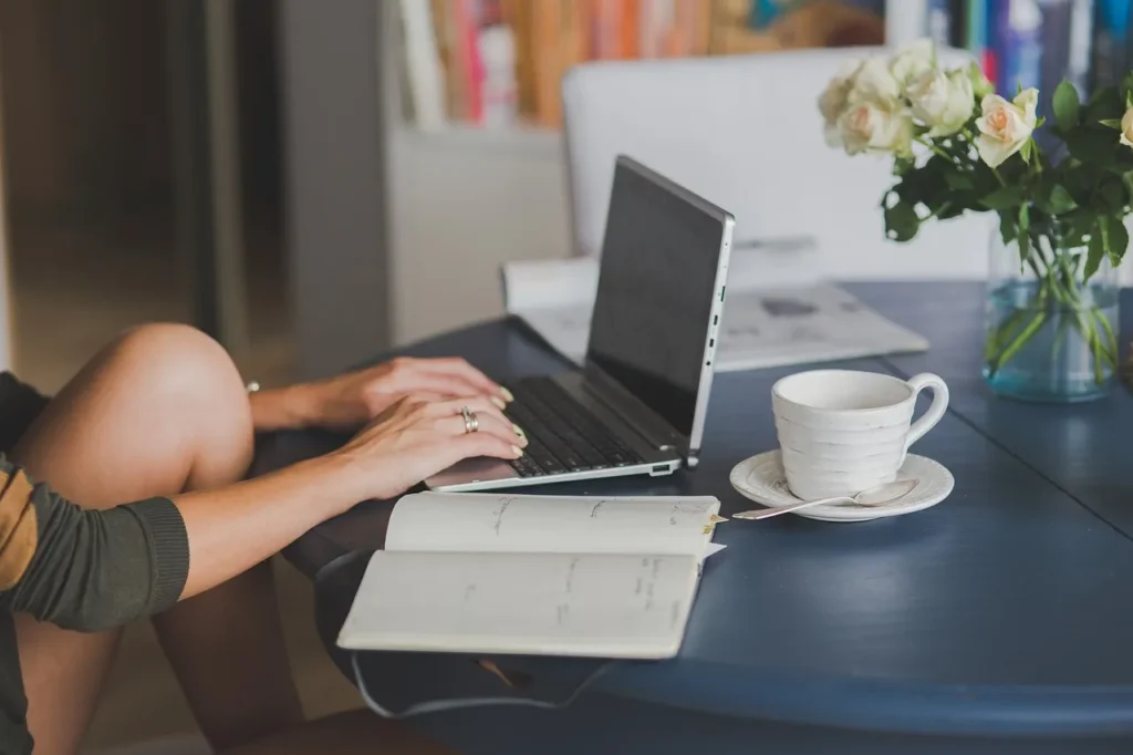 A person's hands typing on a laptop keyboard at a desk with a notebook, a cup of tea, and a vase of flowers.