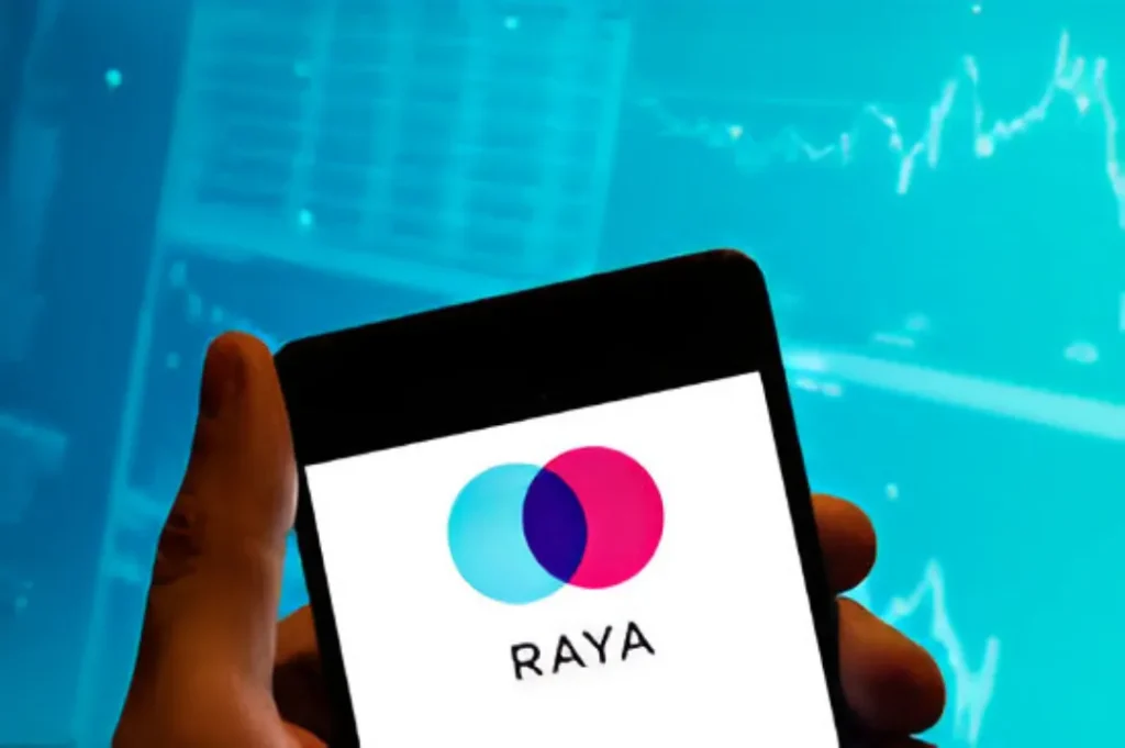 A hand holding a smartphone displaying the Raya dating app logo (overlapping teal and magenta circles) on its screen, with a blurred background showing financial charts in blue tones.