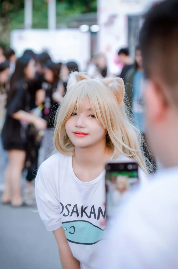 A young woman in a blonde wig with cat ears and green contacts posing for a photo at an anime convention, representing the fan-driven culture of the doujinshi community.