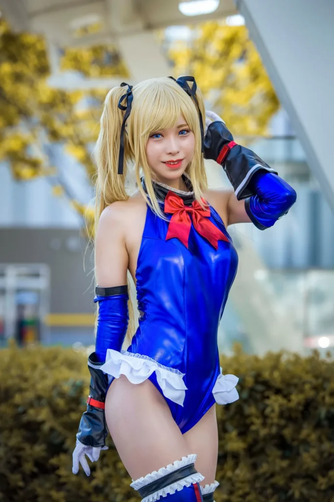 A cosplayer dressed as Marie Rose from Dead or Alive, wearing a signature blue leotard with a red bow, white ruffles, and blonde pigtails, posing outdoors at a convention.