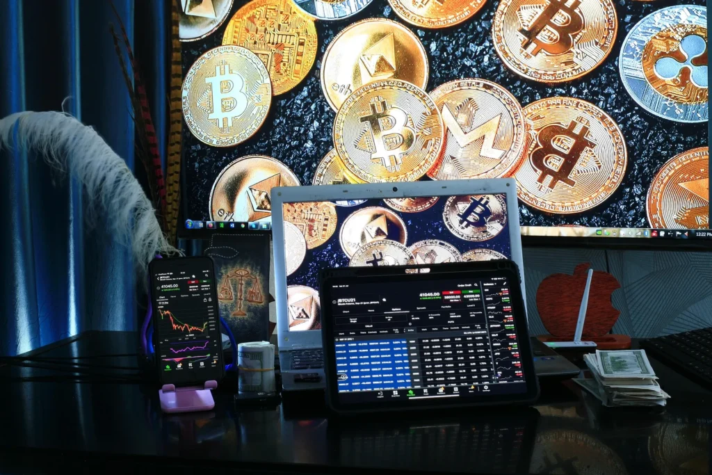 A multi-screen cryptocurrency trading station showing Bitcoin price charts, market data on a tablet, and a background of physical cryptocurrency coins.