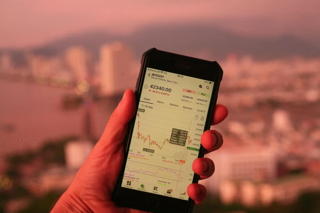 A close-up of a person's hand holding a smartphone displaying a Bitcoin (BTC) price chart and technical indicators with a blurred city skyline in the background.