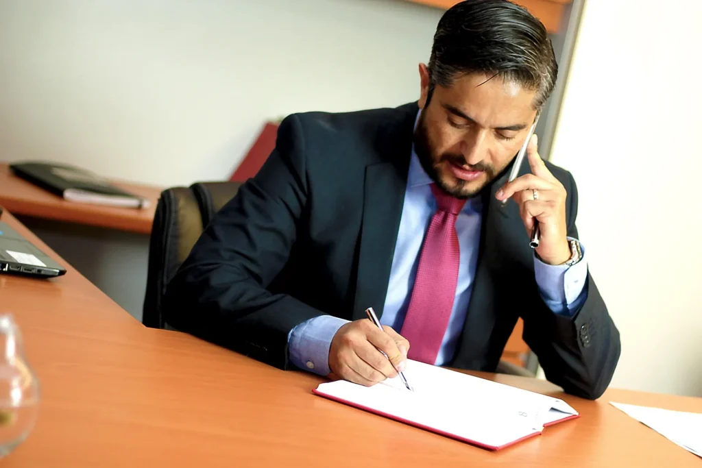 A professional male attorney in a navy suit and red tie sits at a wooden desk, writing in a notebook while speaking on a mobile phone, representing the personalized legal support and debt settlement services offered by Five Lakes Law Group.