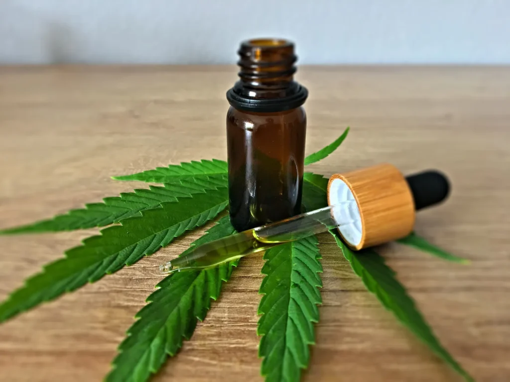 An amber glass bottle of hemp oil sitting on a green cannabis leaf with a glass dropper resting beside it on a wooden surface.