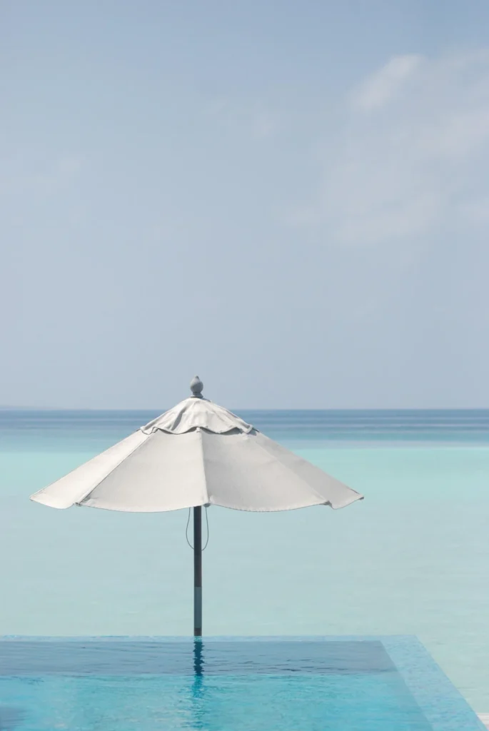 A minimalist view of a white beach umbrella standing by a blue infinity pool, overlooking the turquoise ocean in the Maldives.