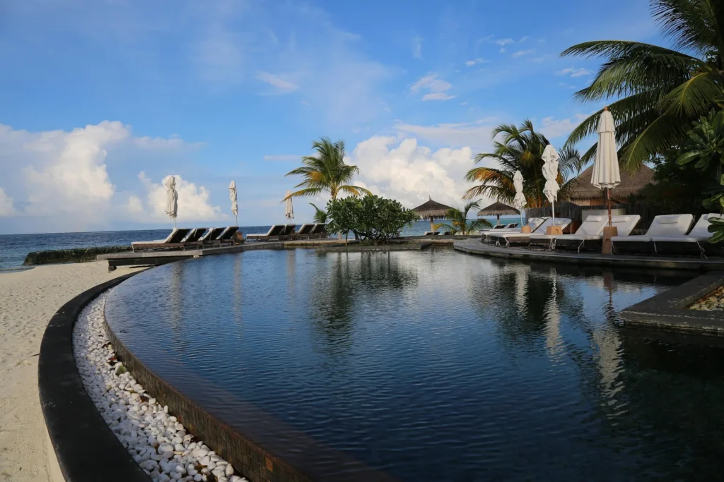 A curved luxury infinity pool lined with white pebbles and sun loungers under palm trees at a tropical beach resort.