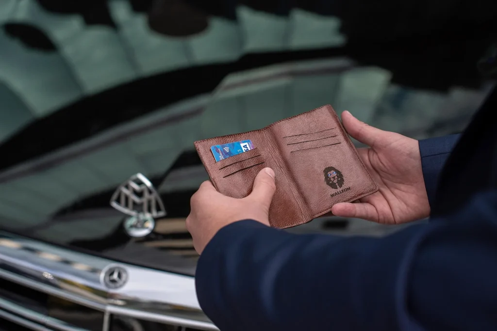A person in a blue suit holding an open brown leather wallet, revealing a credit card and a lion logo, with a luxury Mercedes-Maybach car blurred in the background.