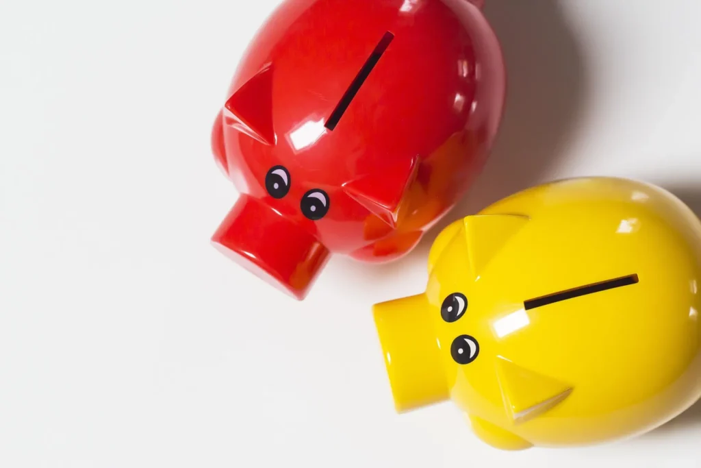Top-down view of two plastic piggy banks, one bright red and one yellow, sitting side-by-side on a clean white surface.