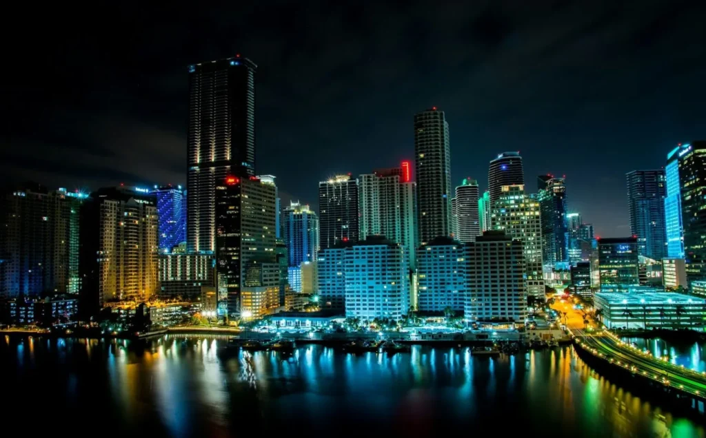 A vibrant night photo of the modern Miami skyline from across the bay, with high-rise buildings illuminated with blue, cyan, and warm lights. The colorful city lights and building façades are vividly reflected on the dark water surface in the foreground, with an elevated roadway and marina visible. The scene captures the dense urban and financial character of Miami.
